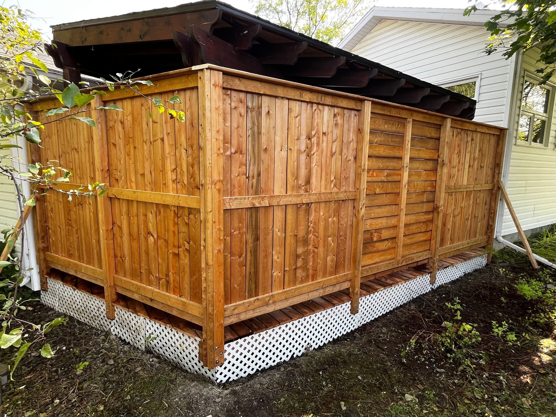 Clôture en bois brun avec toit noir, adossée à un bâtiment. Socle en treillis blanc.