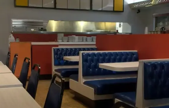 Restaurant booths with blue vinyl seats and small white tables, orange and gray walls, kitchen visible in background.