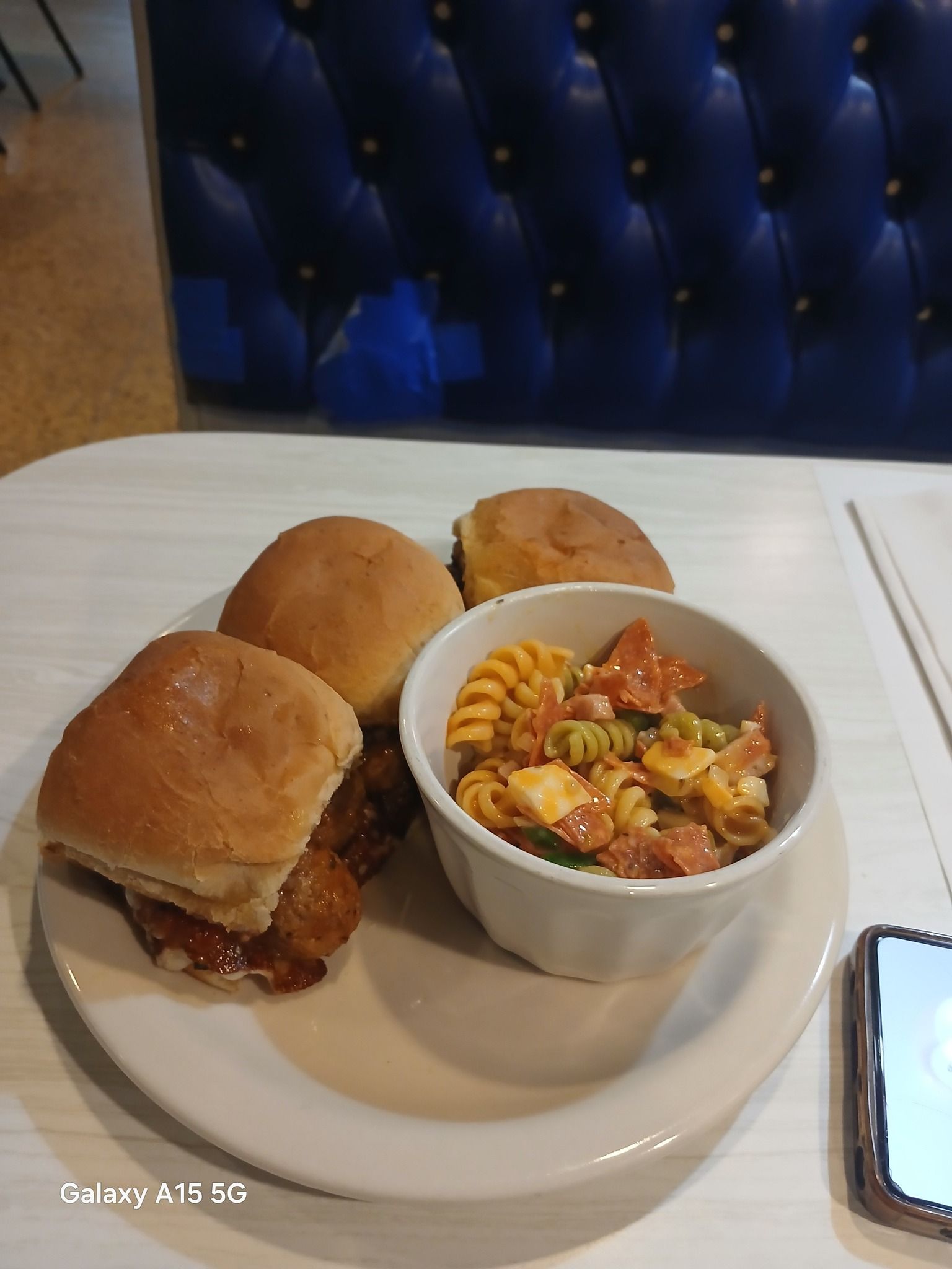Plate of food: burger sliders, pasta salad, white plate, white table, blue booth.