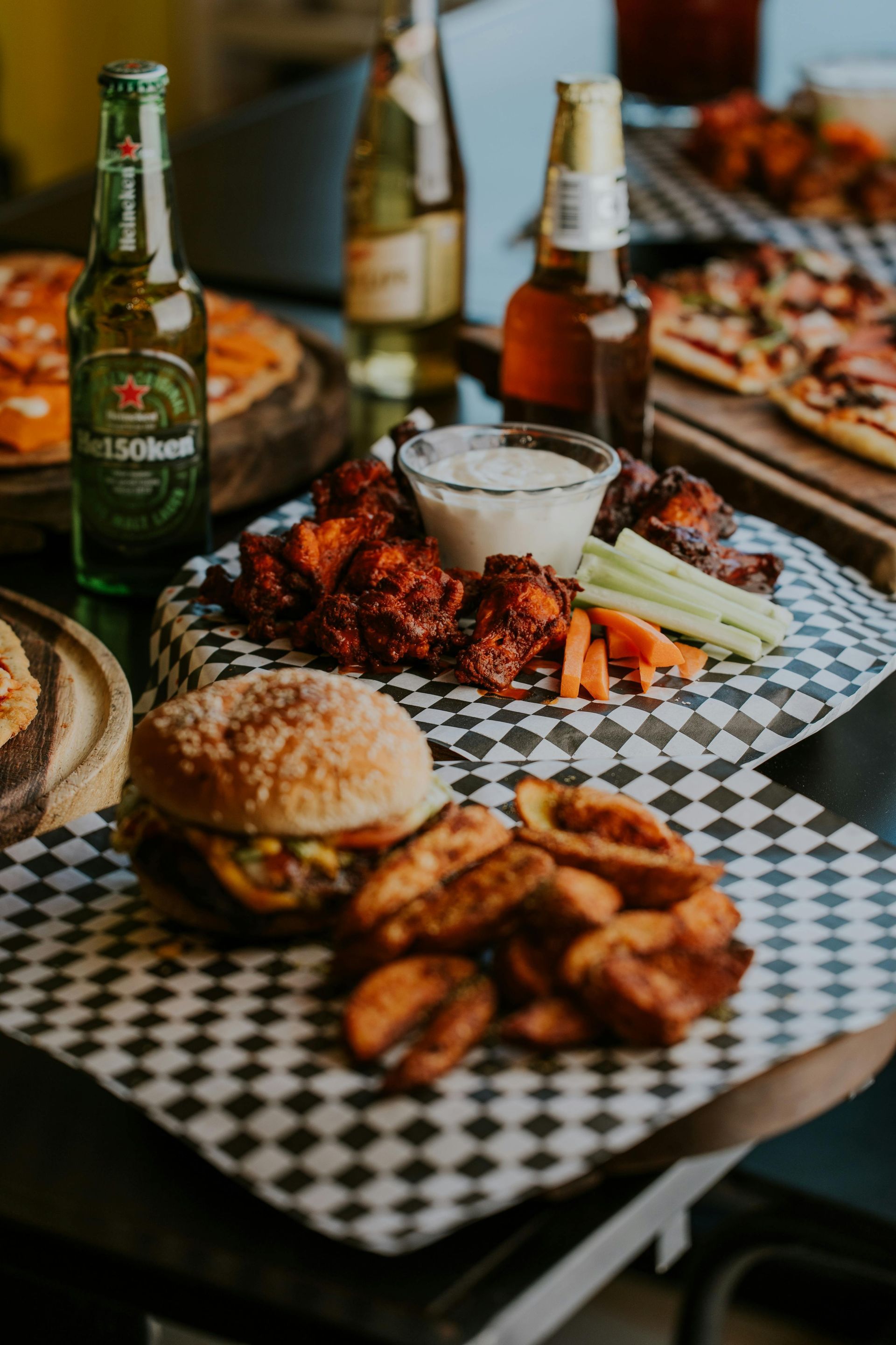 Food spread: burger, wings, wedges, pizza, and beer bottles on checkered paper.