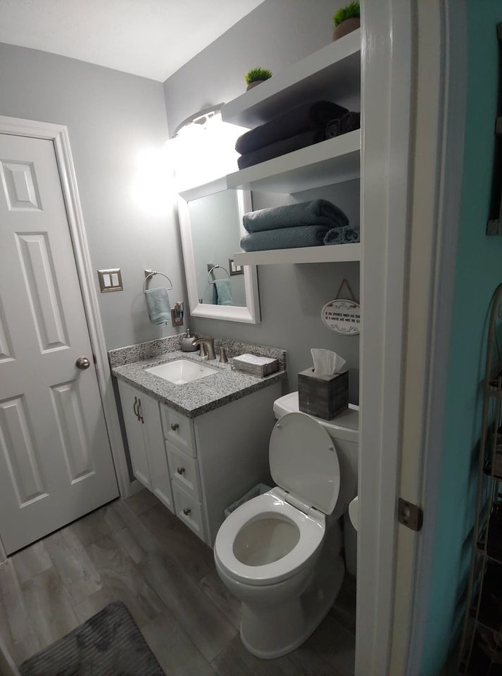 A bathroom with a toilet , sink , mirror and shelves.