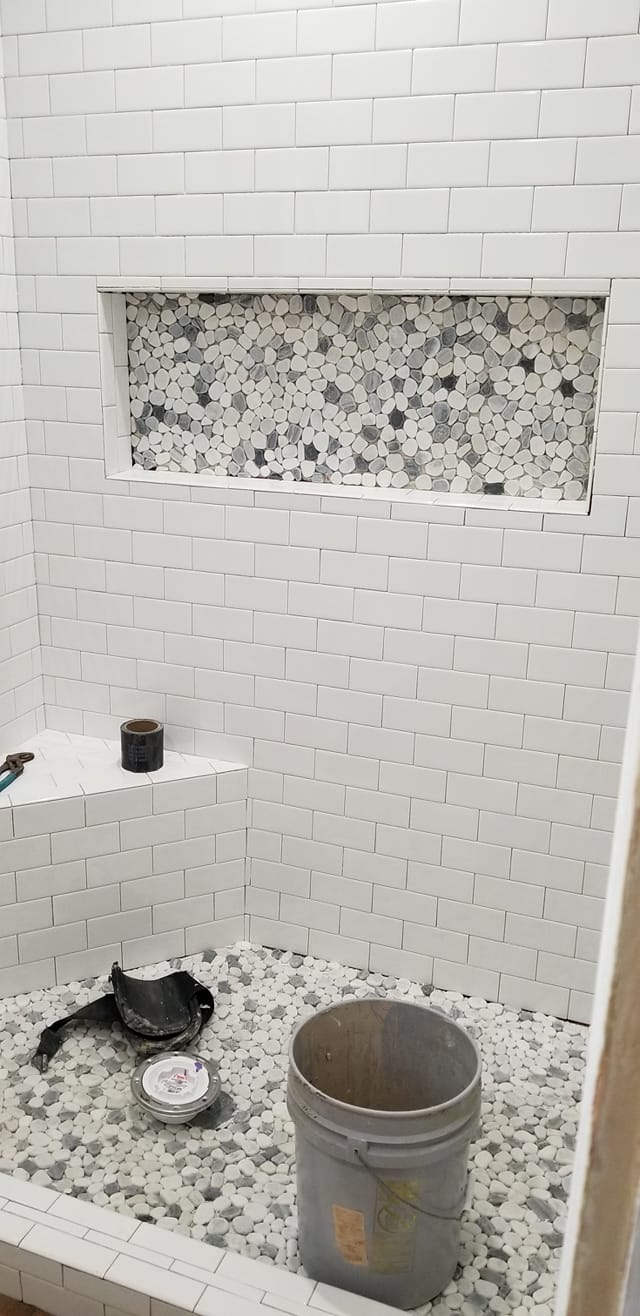 A shower with white tiles and a bucket on the floor.