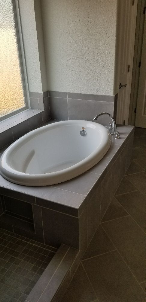 A white bathtub is sitting on top of a tiled counter in a bathroom next to a window.
