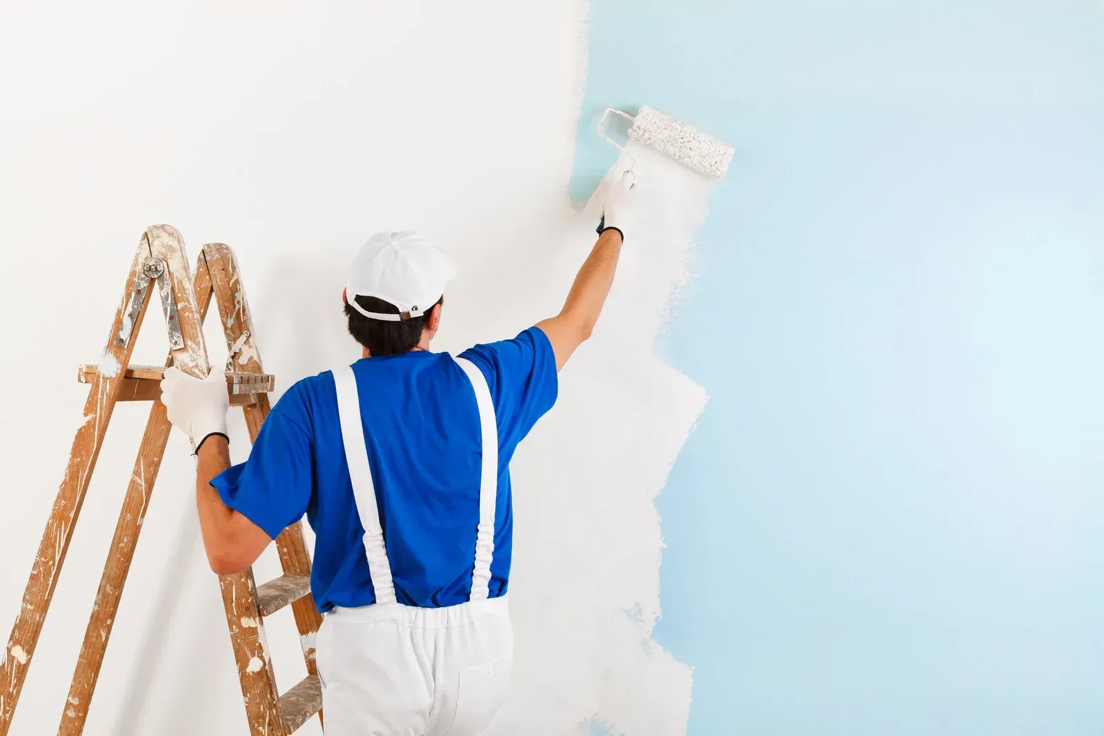 Painter in blue overalls and white cap painting a light blue wall next to a ladder