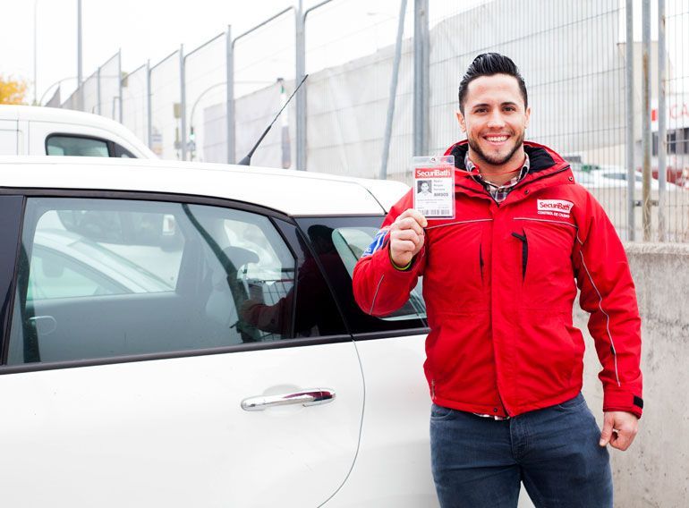 Un hombre con una chaqueta roja sostiene una licencia de conducir frente a un automóvil blanco.