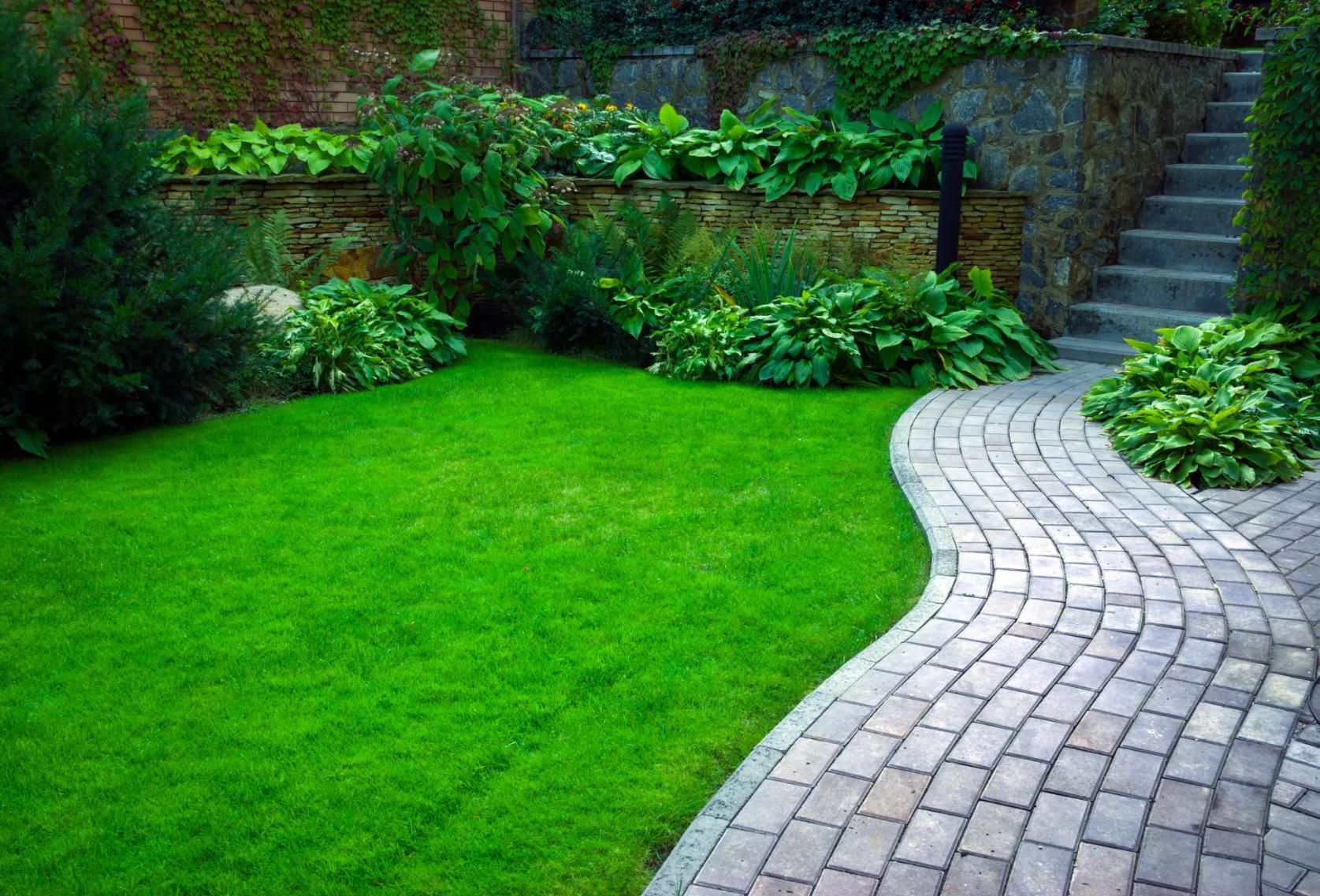 Green lawn with a winding brick path, bordered by shrubs and a stone wall with steps.