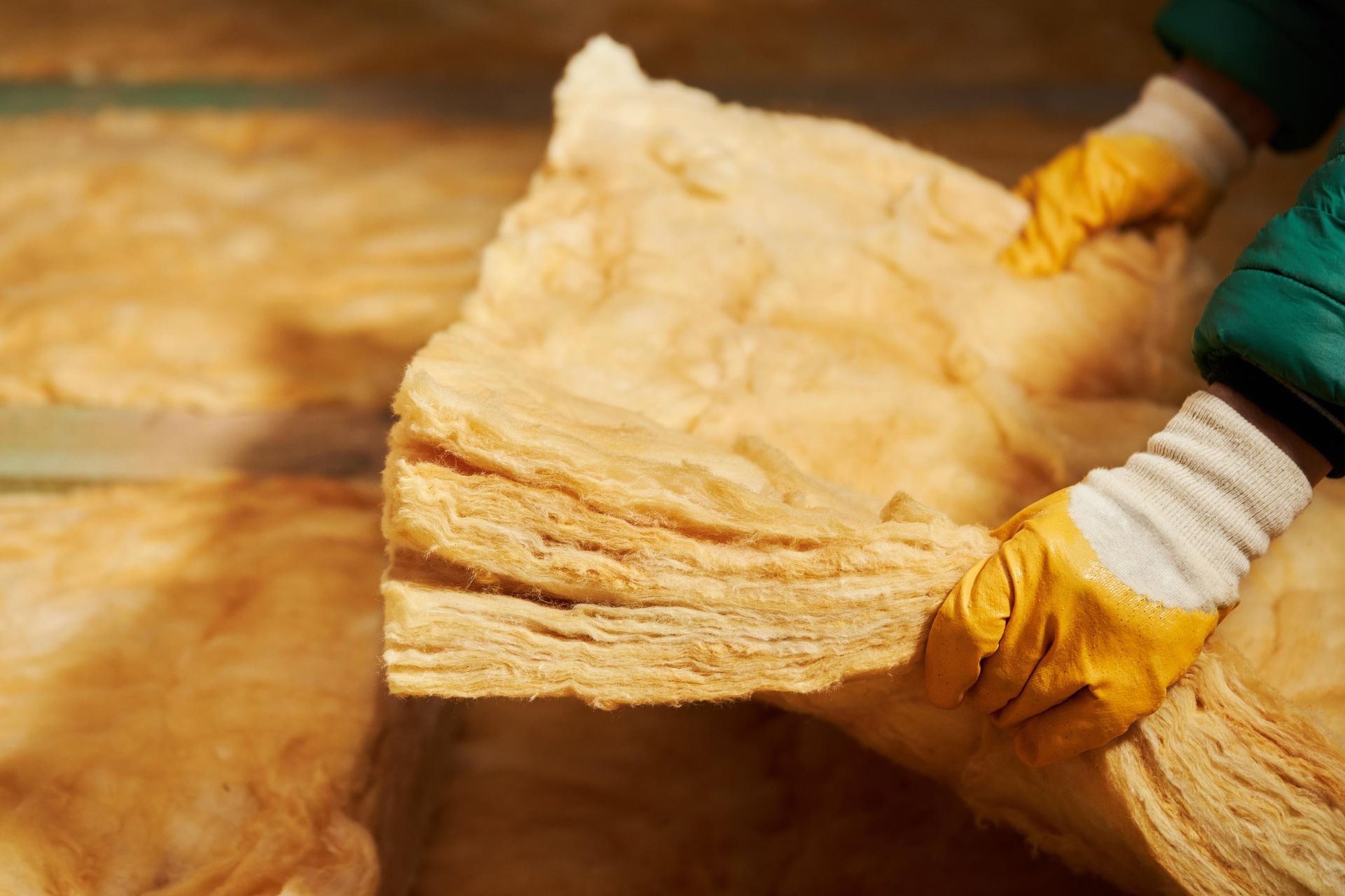 Hands in yellow gloves holding insulation, with a stack of insulation in the background.