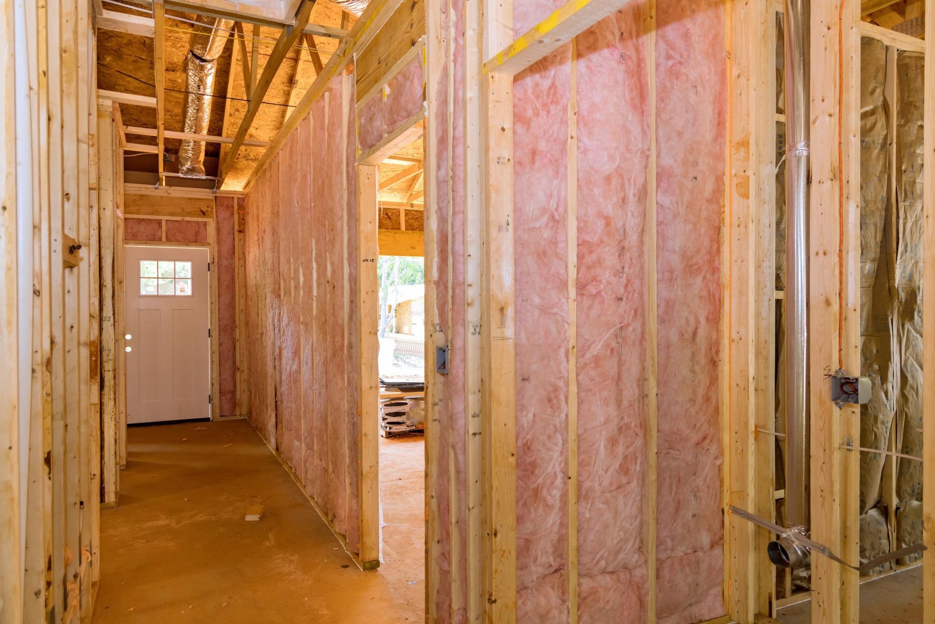 Interior view of a building under construction, showing wooden framing, pink insulation, and a hallway with a door.
