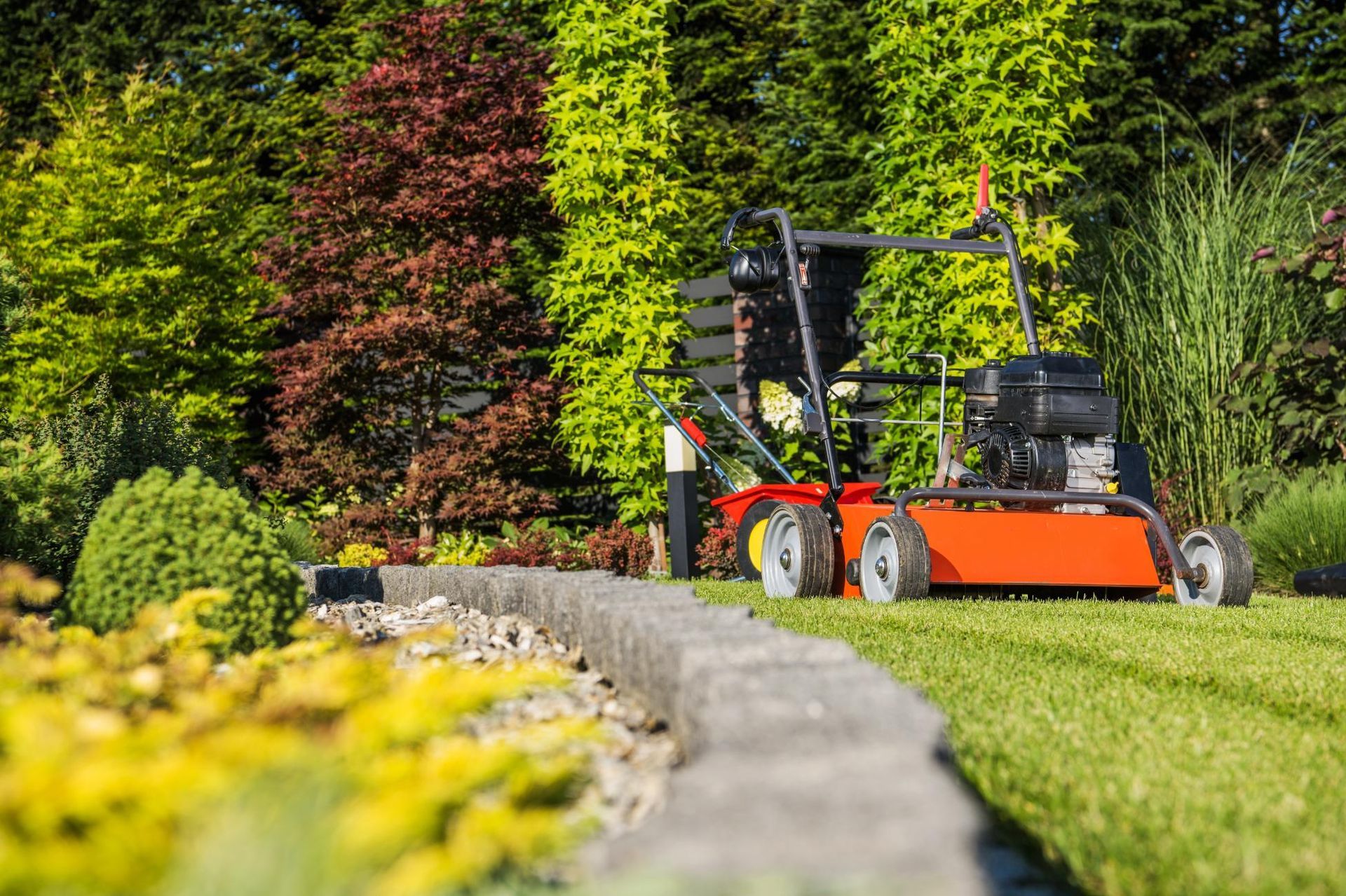 Lawn aerator on green grass near a stone border, with colorful trees and plants in the background.