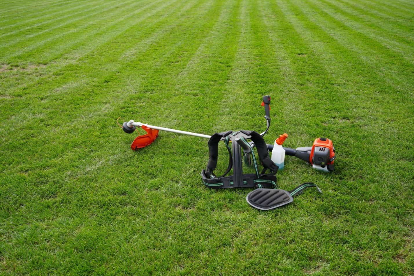 A gas-powered backpack string trimmer rests on a green, striped lawn.