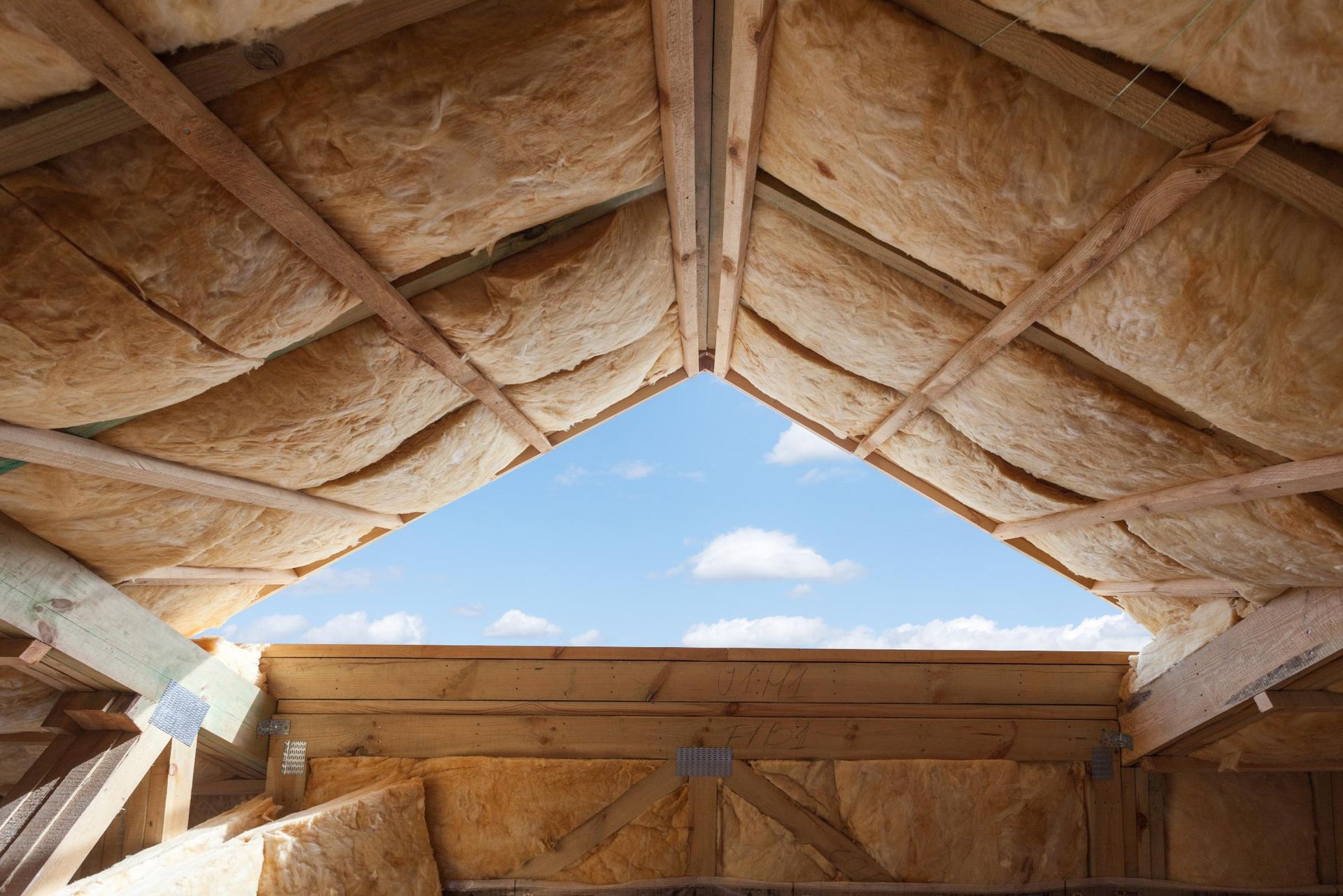 Interior view of an unfinished roof, insulation visible, with a triangular opening to the blue sky.