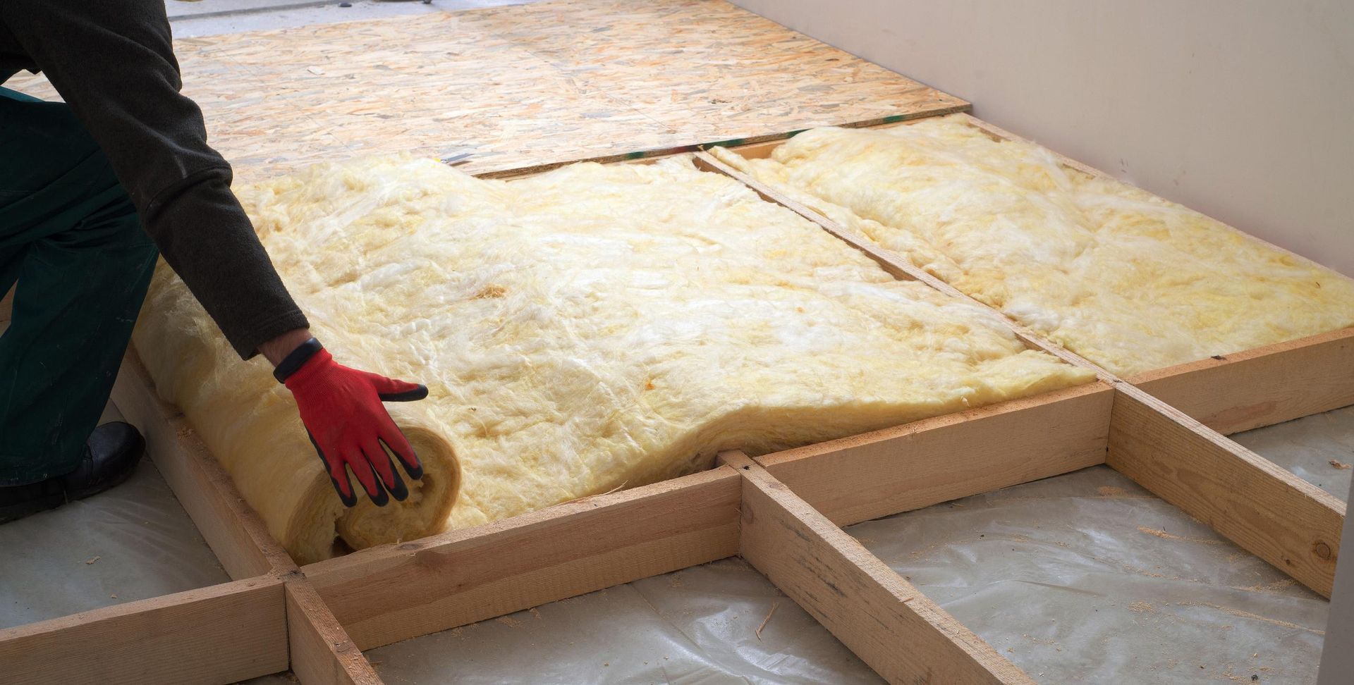 Person installing insulation between wooden floor joists; red-gloved hands unrolling yellow insulation.