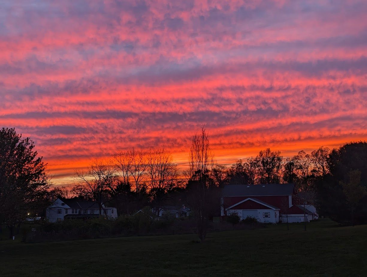 A sunset over a field with a house in the foreground and trees in the background.