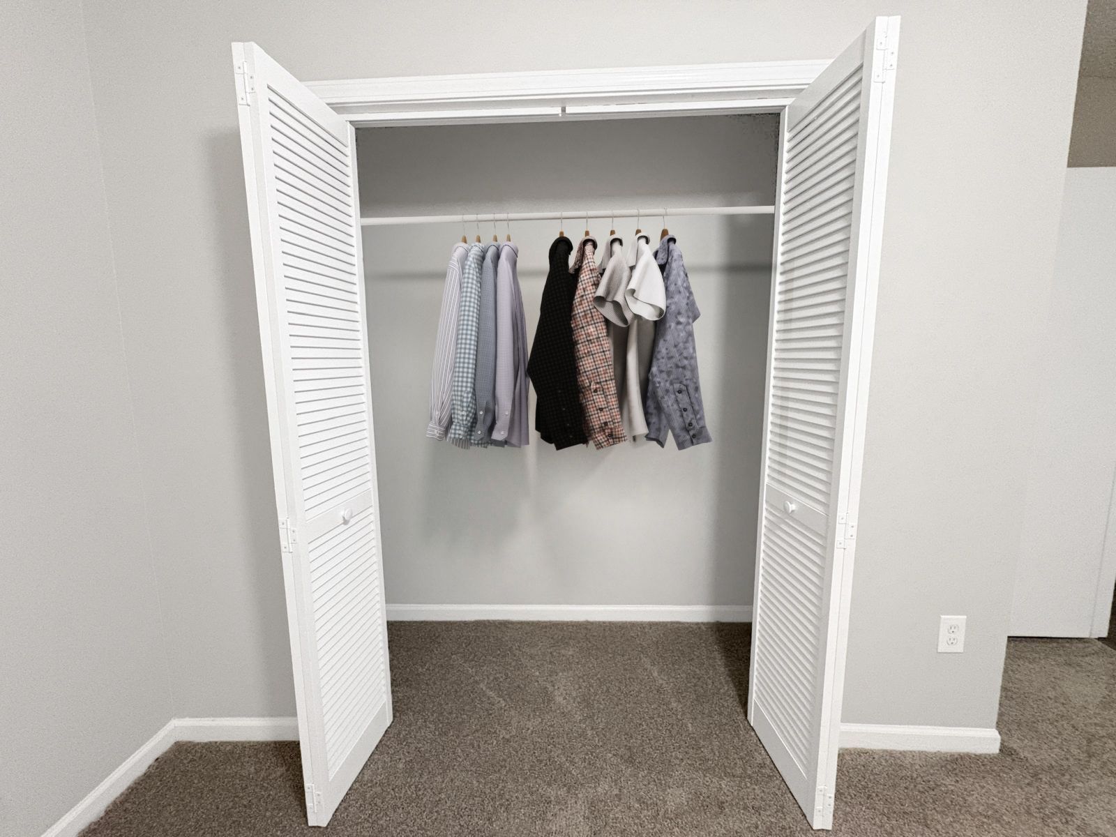 Open closet with white louvered doors, carpeted floor, and clothes hanging inside.