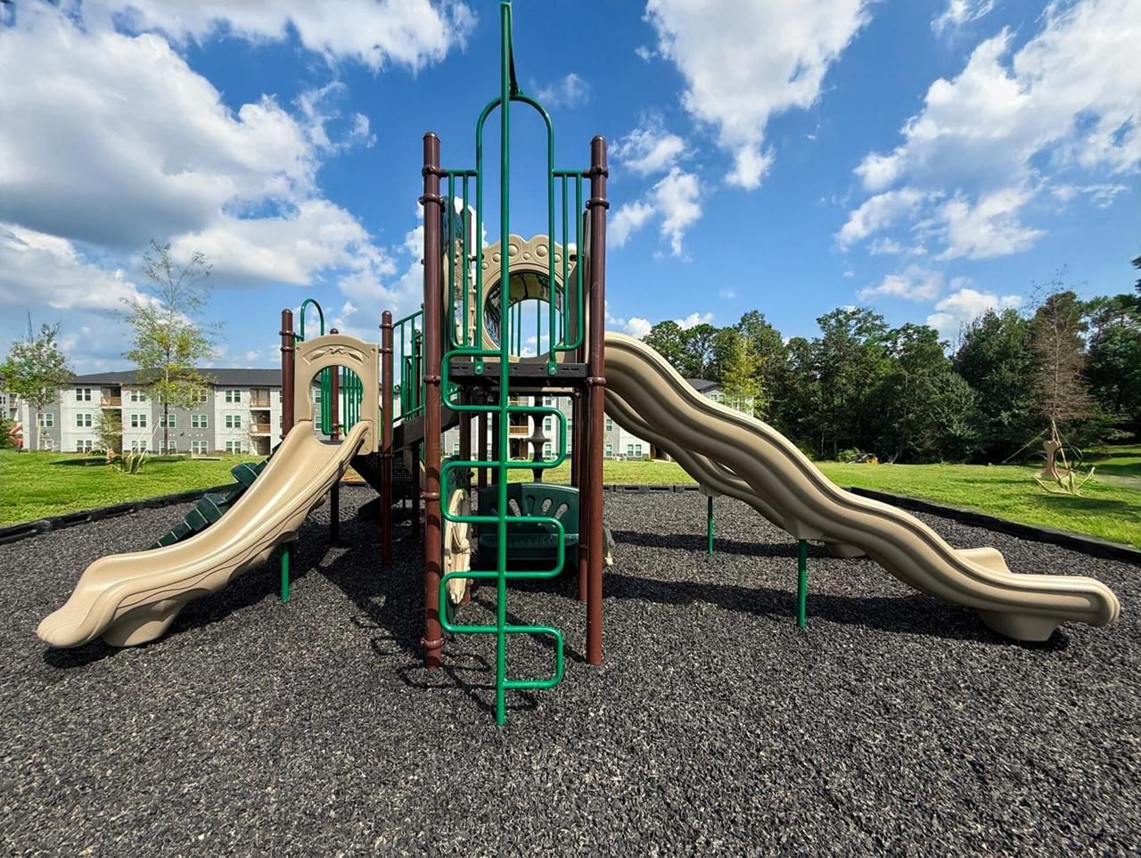Playground with tan slides, green climbing frame, and gravel ground under a blue sky
