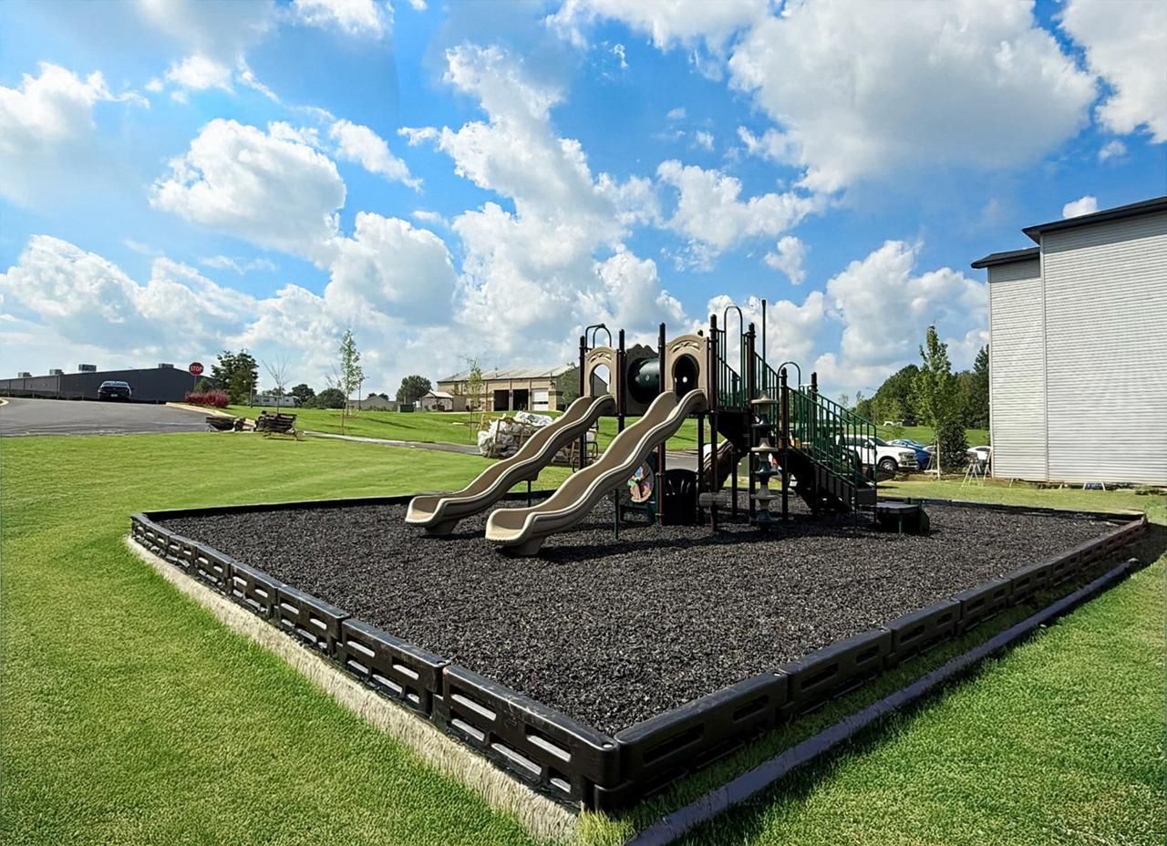 Two playground slides on a black rubber play area under a blue sky with clouds, beside a building and grassy field