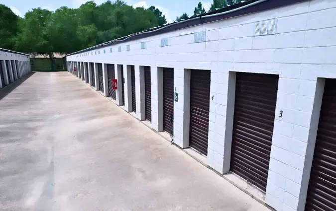 Rows of brown storage unit doors on a white building, concrete ground, green trees in background.