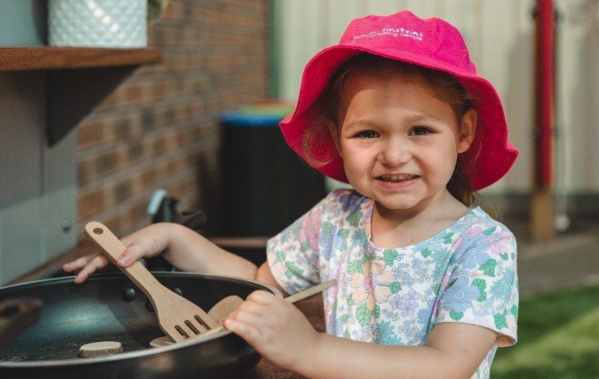A little girl wearing a pink hat is holding a wooden spatula over a pan.