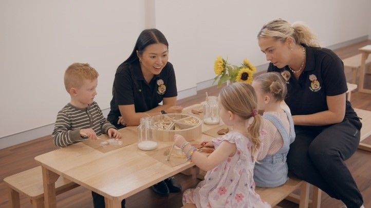 A group of children are sitting at a table with two teachers.