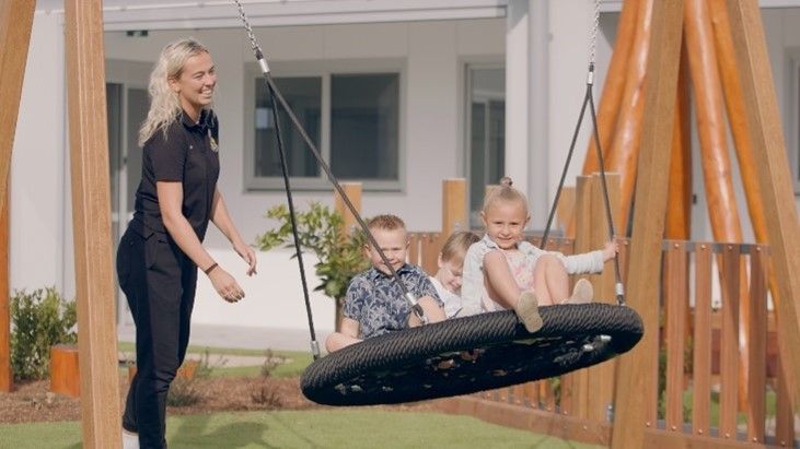A woman is standing next to two children sitting on a tire swing.