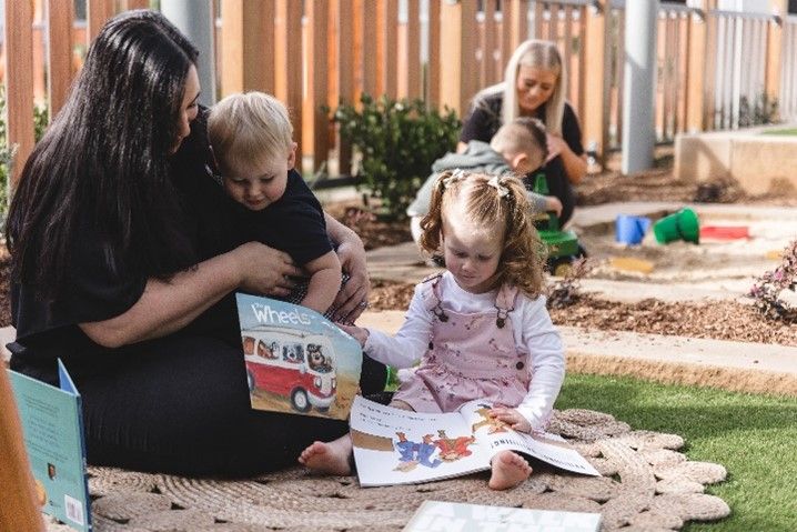 A woman is sitting on the ground reading a book to two children.