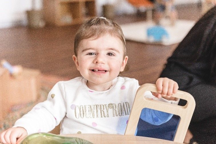 A little girl is sitting at a table holding a mirror and smiling.
