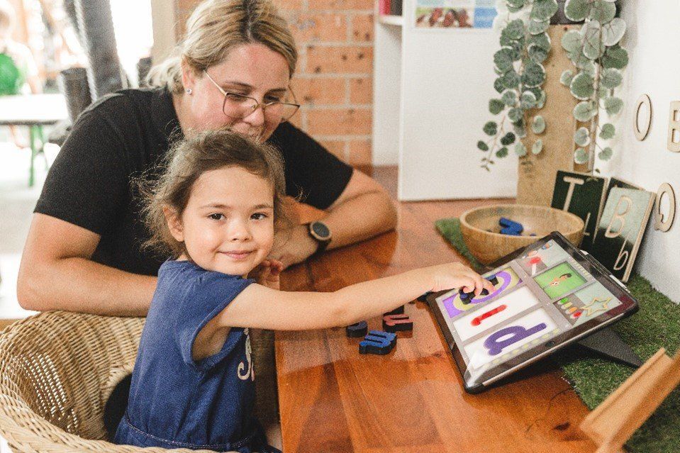 A woman and a little girl are sitting at a table playing with a tablet.
