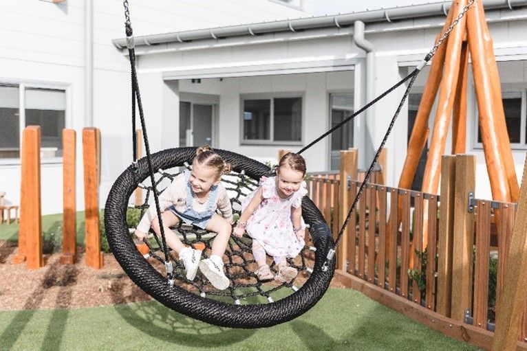Two little girls are sitting on a swing in a playground.