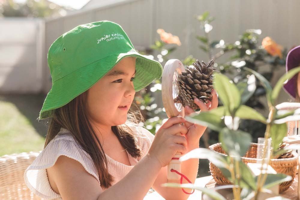 A little girl in a green hat is looking at a pine cone through a magnifying glass.