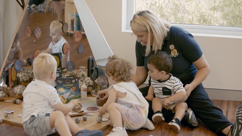 A teacher assists three young children playing with toys inside a brightly lit room, near a decorative teepee.