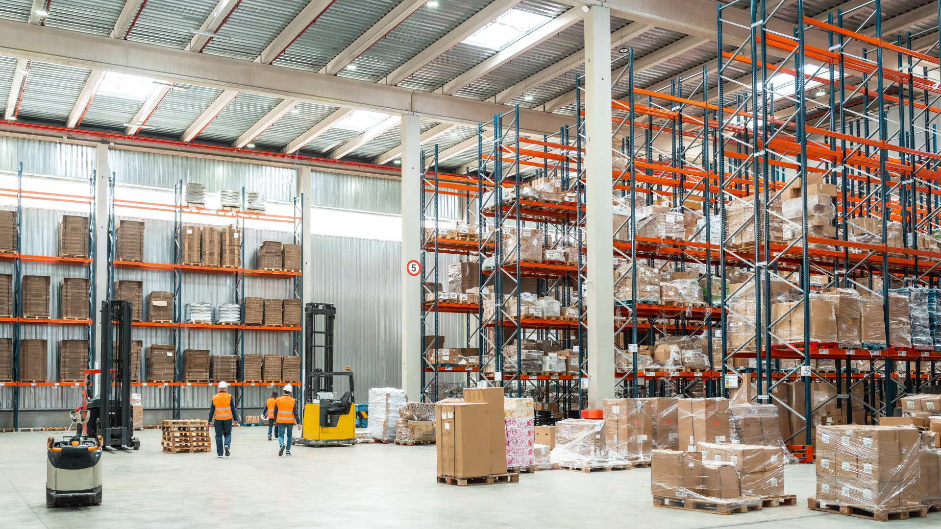 Warehouse interior with workers, forklifts, and stacked pallets on tall orange storage racks