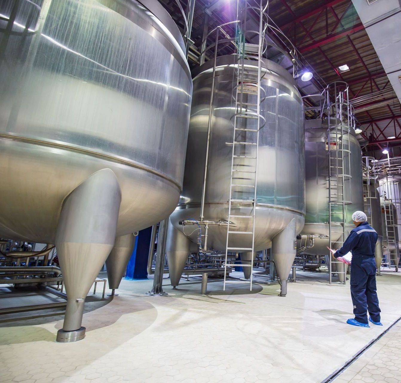 Worker in blue overalls inspecting large stainless steel tanks in a factory setting.