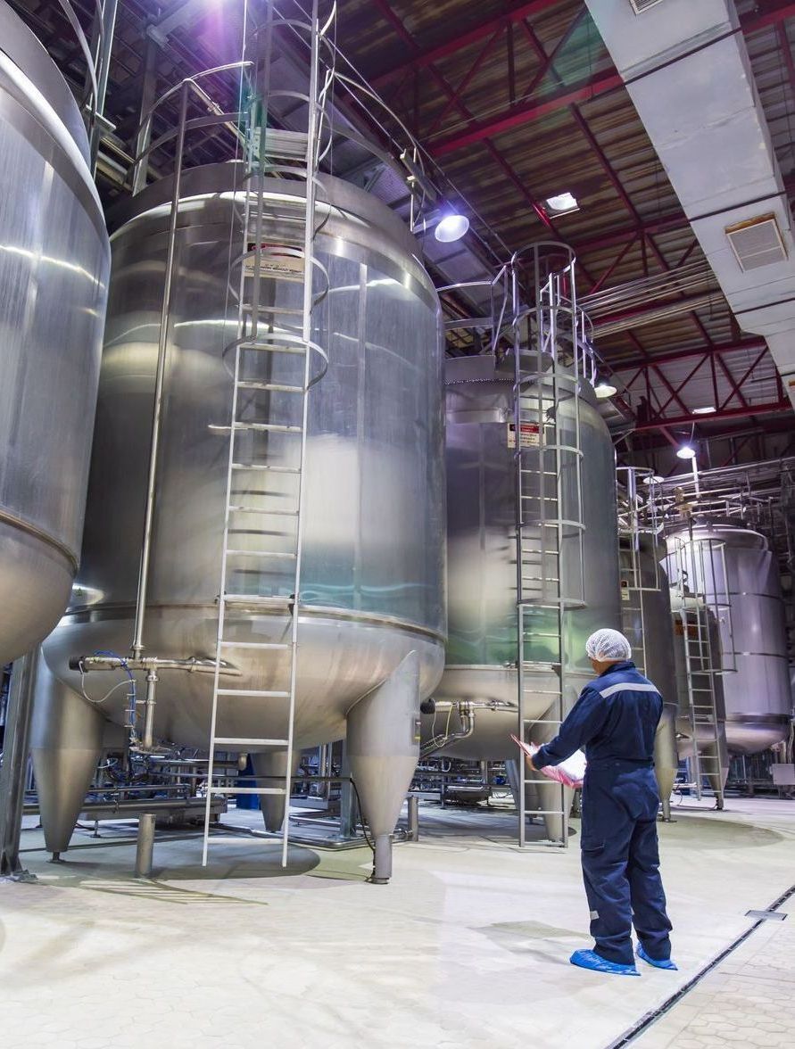 A man is standing in front of a row of stainless steel tanks in a factory.