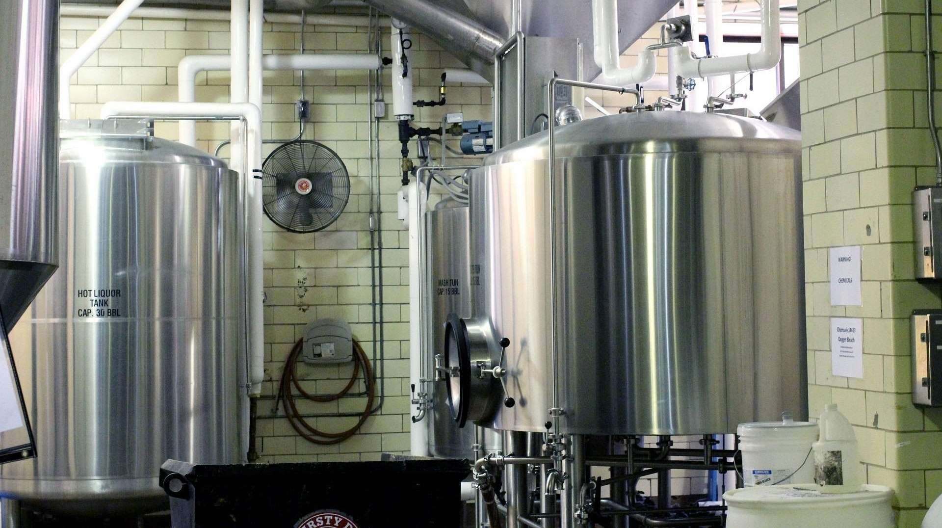 Stainless steel brewery equipment inside a brick-walled industrial space; a fan and pipes are visible.