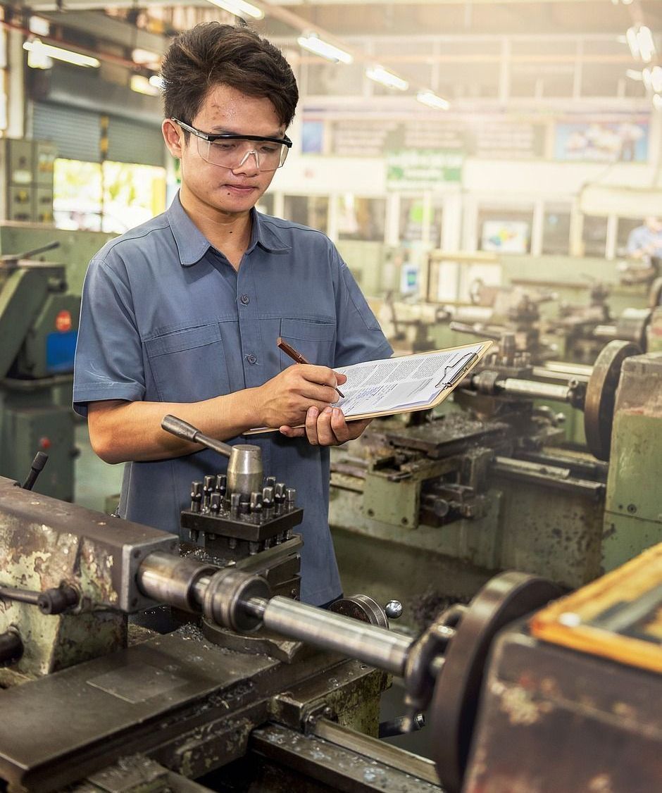 Man in safety glasses taking notes in a machine shop, standing near a lathe.