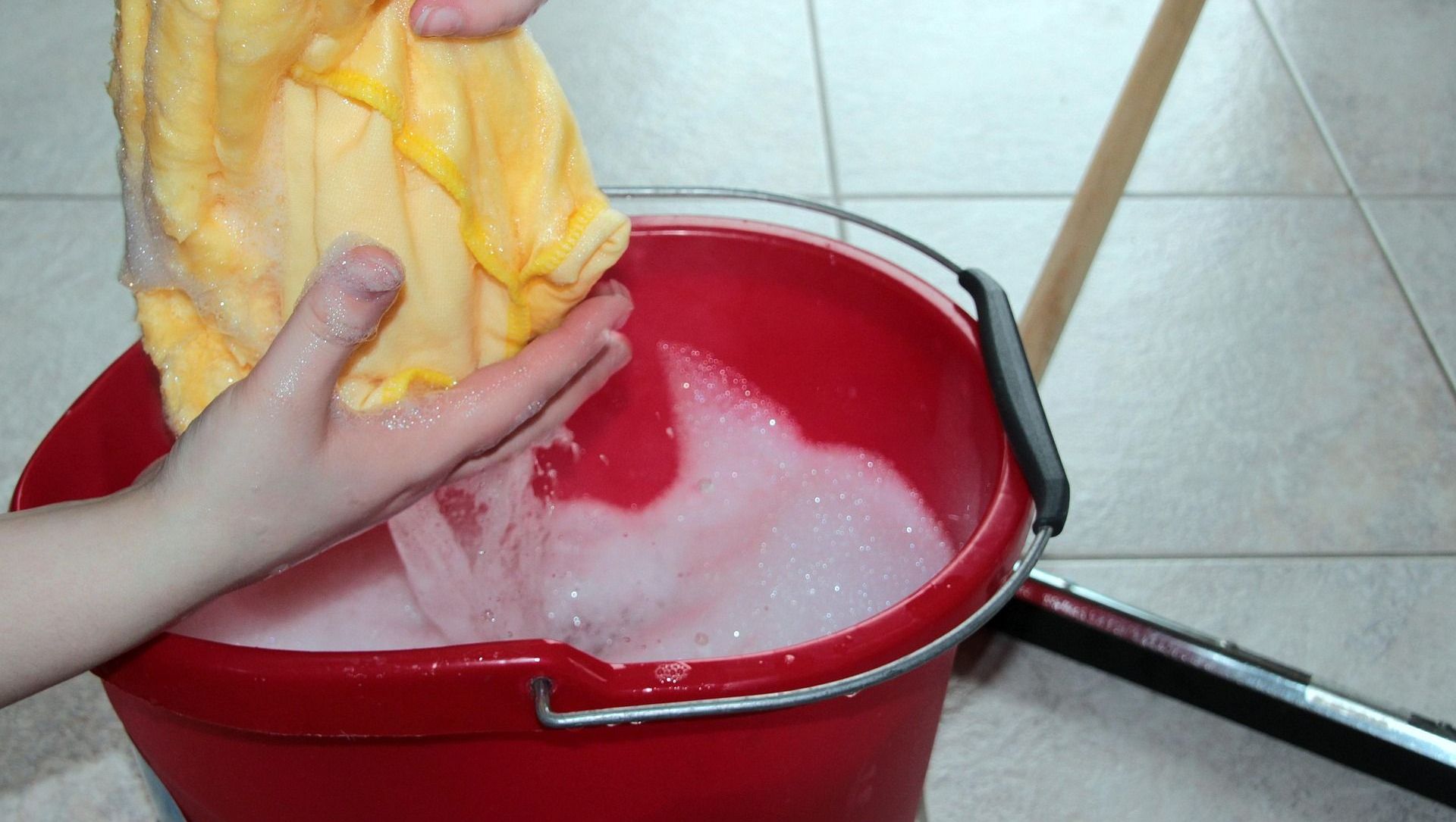 A person is holding a yellow cloth over a red bucket of soapy water.