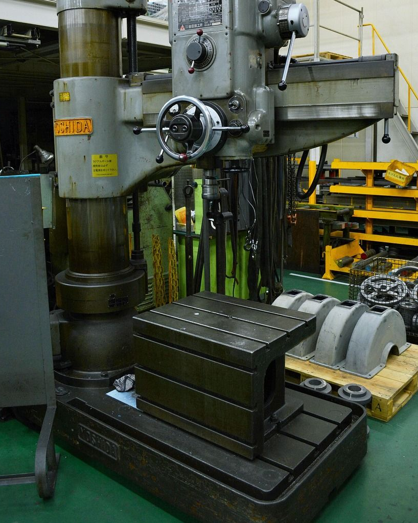 Radial drill press with a metal workpiece on its base in a workshop.