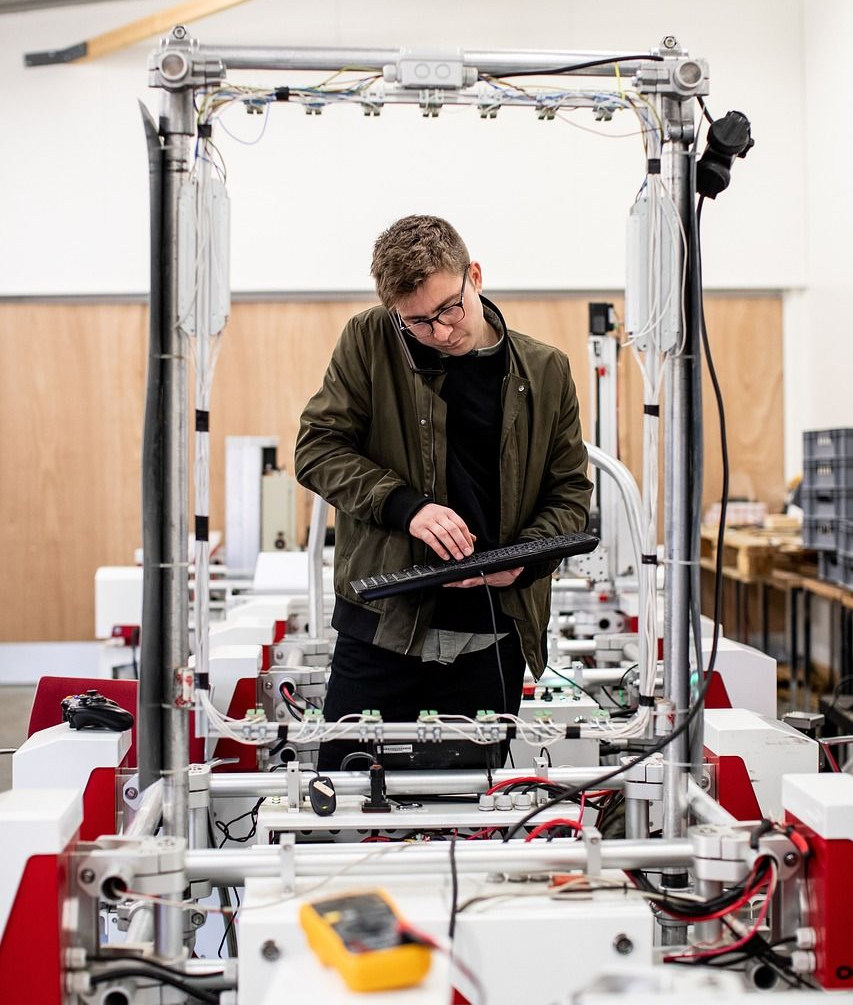 Man working on machinery while talking on the phone; wearing glasses, bomber jacket; industrial setting.
