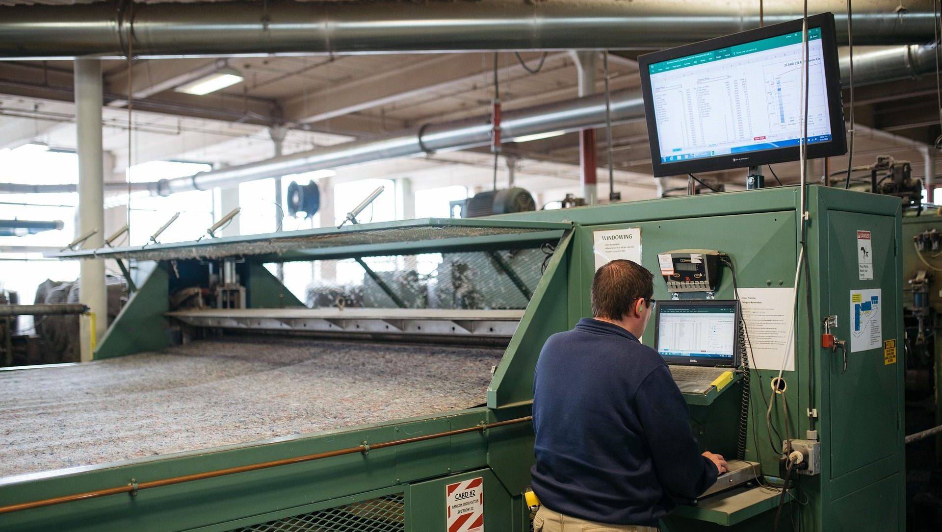 Person operating machinery in a factory; monitoring a screen; large machine in the foreground.