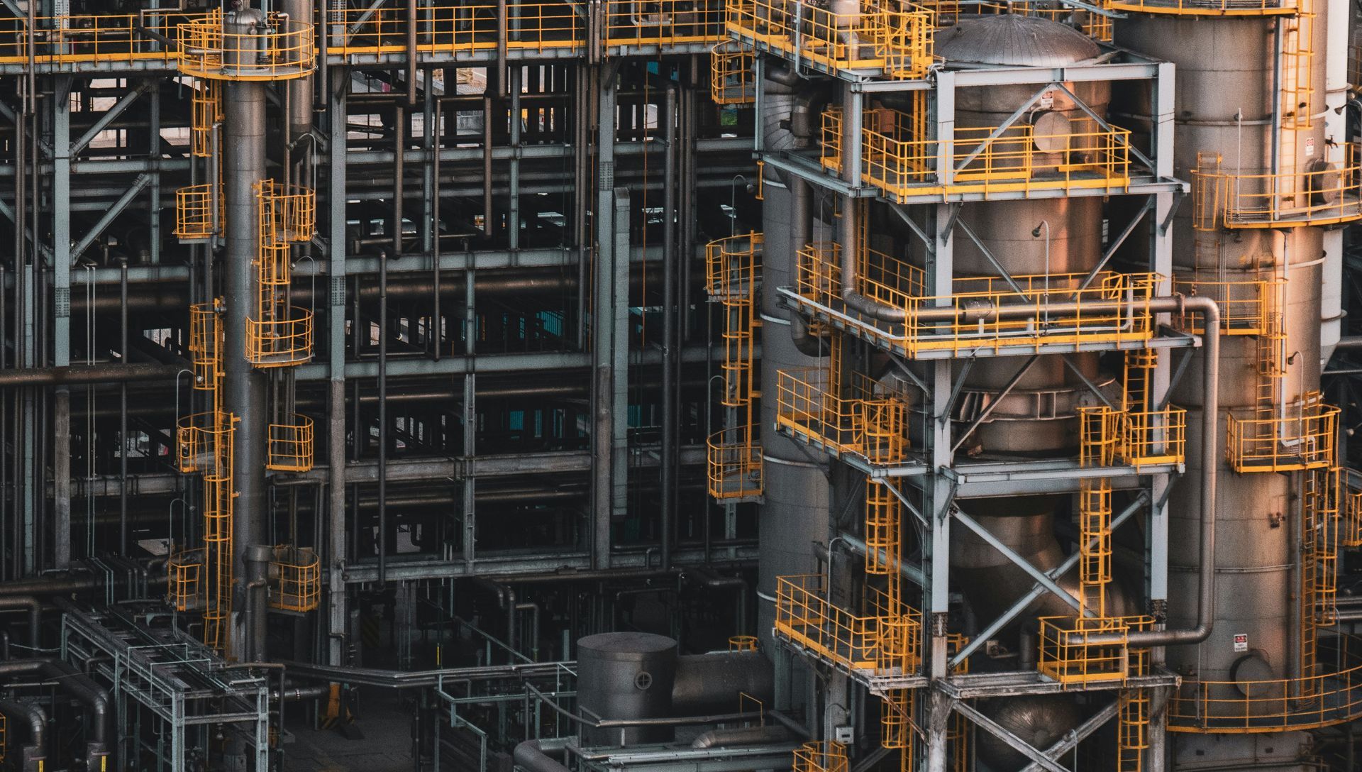 Industrial plant with towers, pipes, and scaffolding, mostly gray with yellow accents.