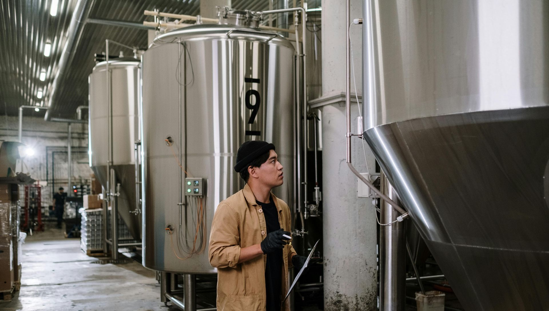 Man in a brewery inspecting stainless steel tanks, holding a clipboard.