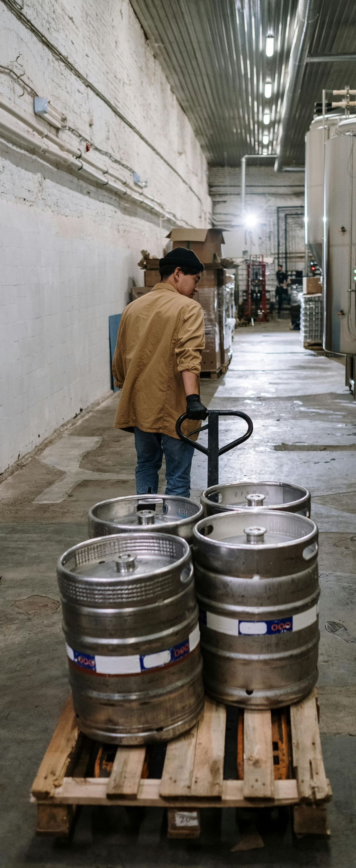 A person transporting beer kegs on a pallet with a hand truck in a warehouse.