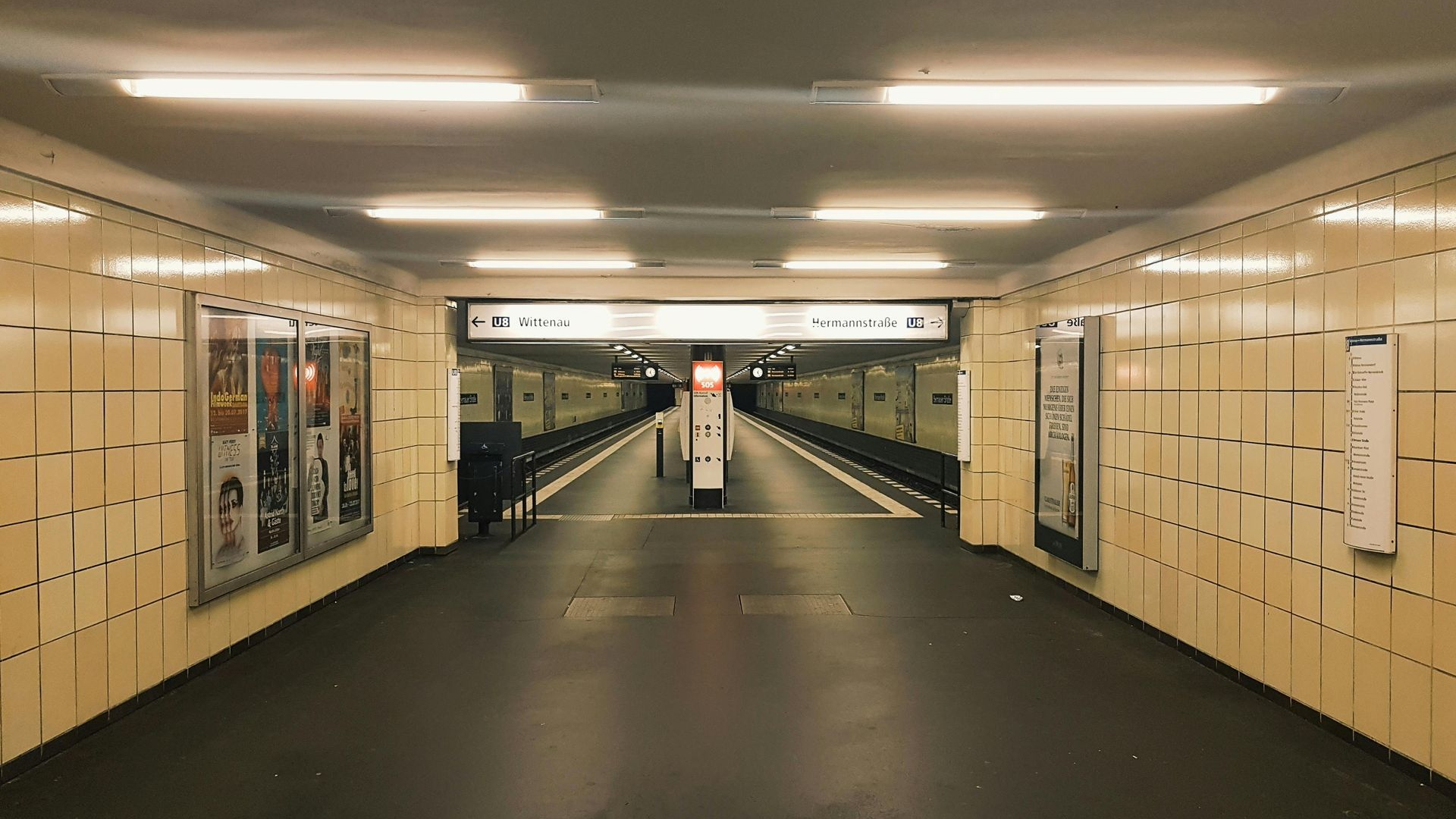 Subway station platform with tiled walls and overhead fluorescent lights.