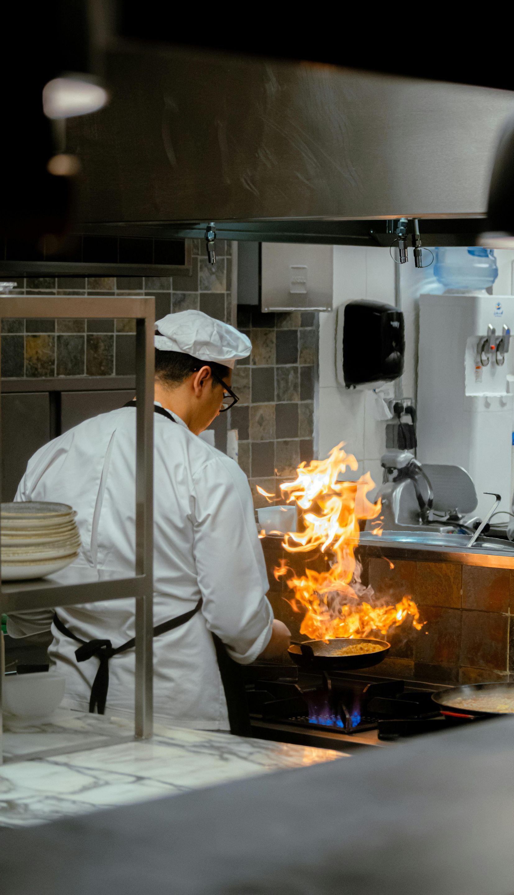 A chef is cooking food in a kitchen with flames coming out of a pan.