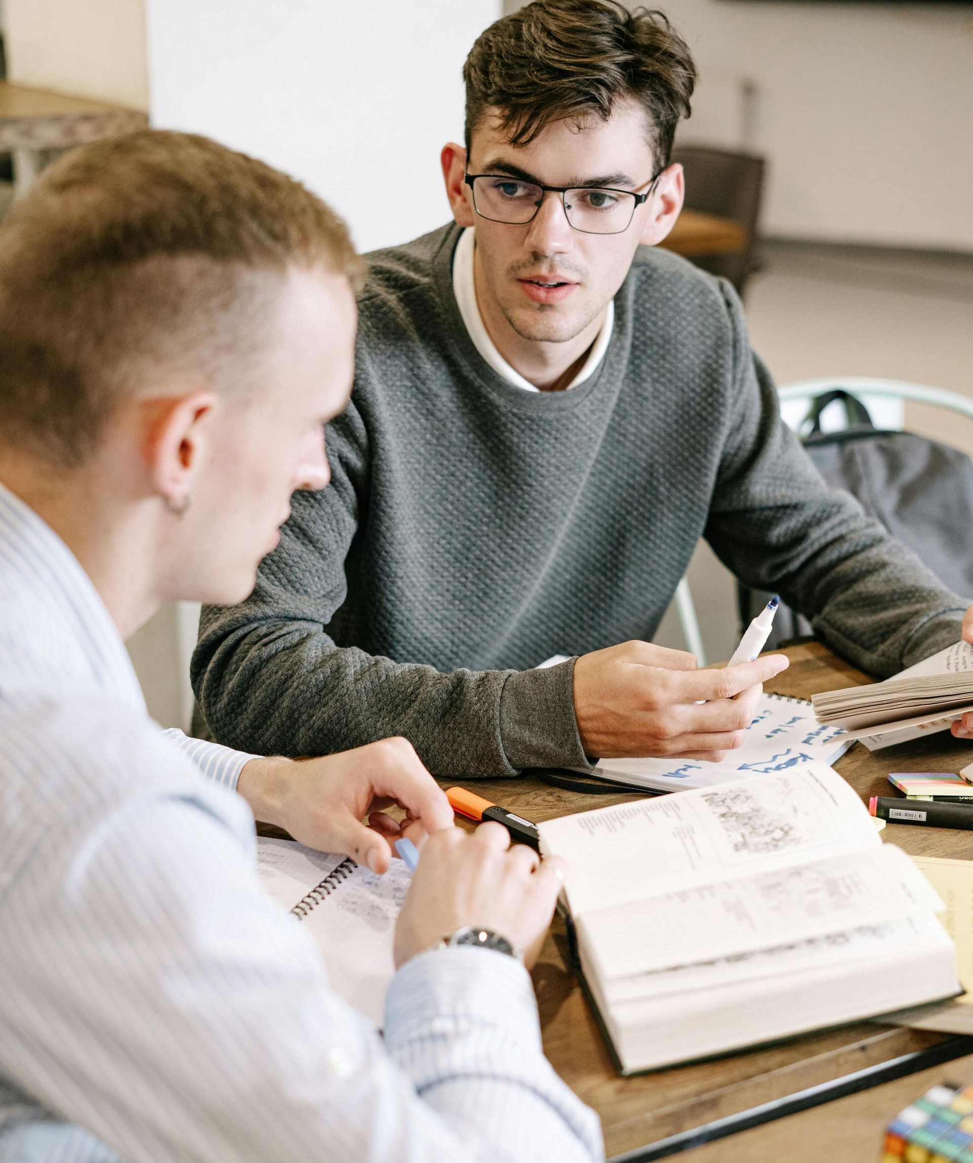 Two men sit at a table, looking at documents. One man in a gray sweater points, while the other in a light blue shirt looks on.