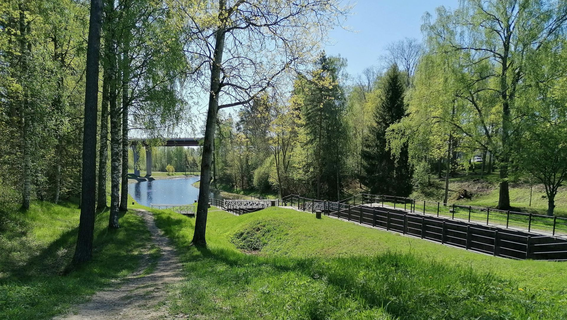 Lush green park with trees, grass, and a water body. A bridge is visible in the distance. Sunny day.