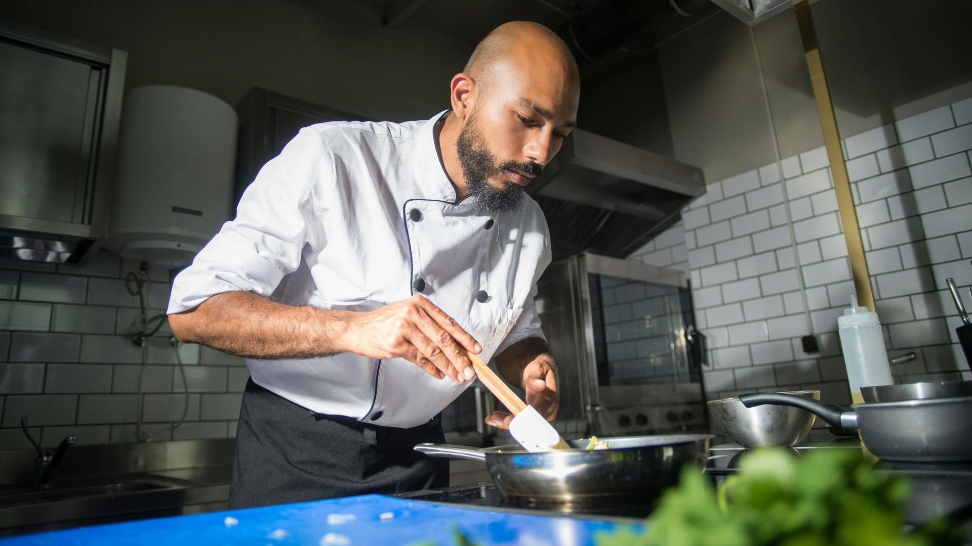 A chef is stirring food in a pan with a wooden spoon in a kitchen.