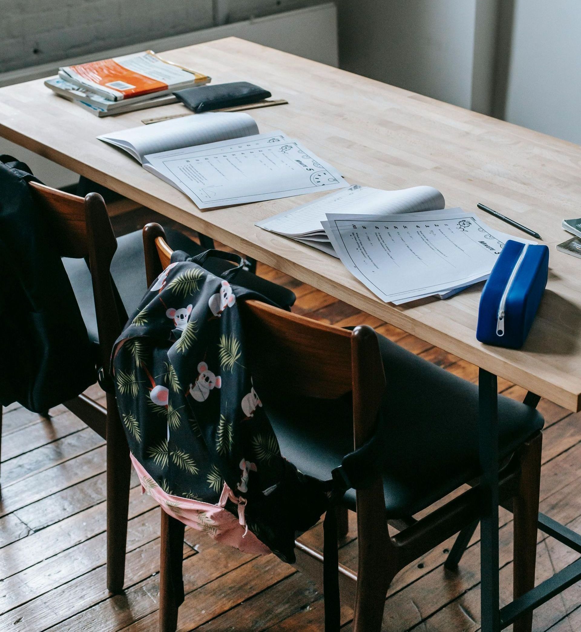 Wooden table with papers, books, and pencil case, two chairs with a floral scarf draped over one, on a wooden floor.