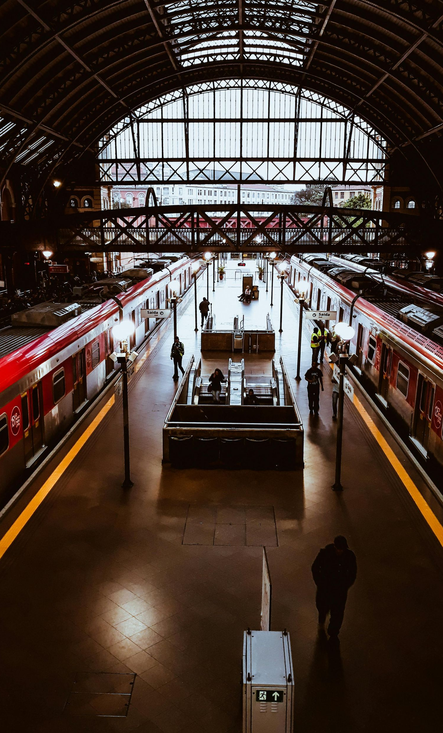 Train station interior with trains at platforms, people walking, overhead glass roof.