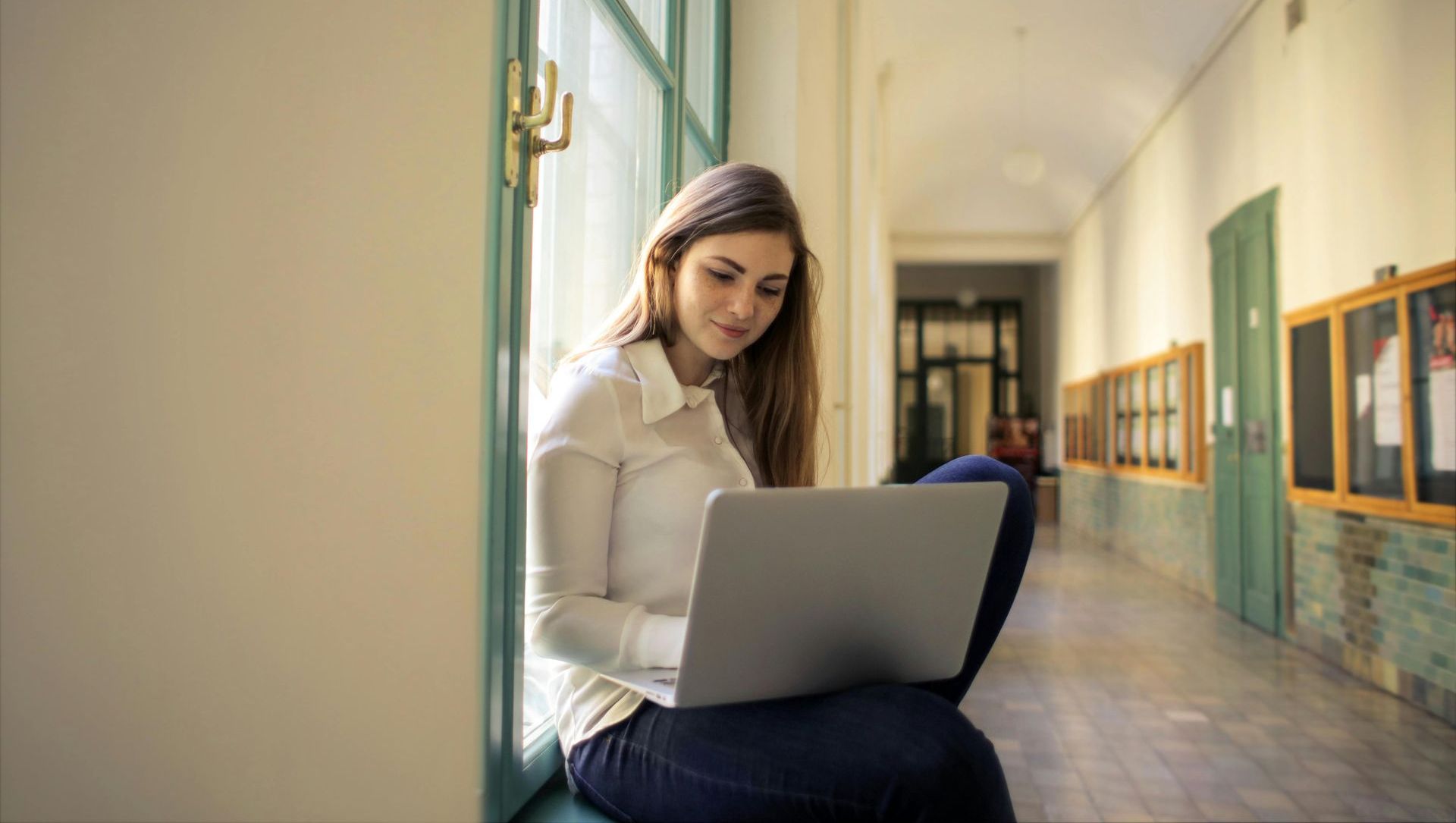 Woman with long brown hair sits on a window ledge, using a laptop in a brightly lit hallway.